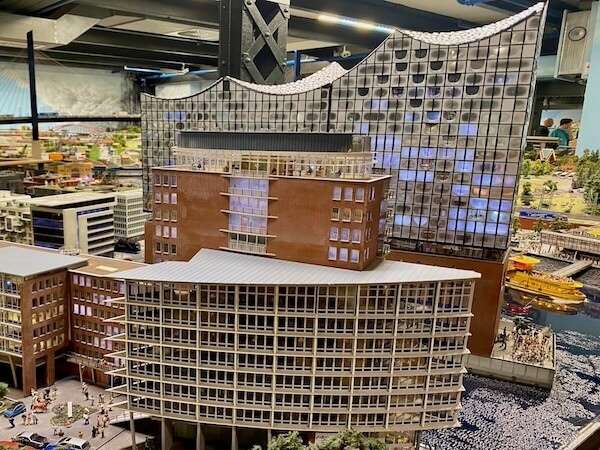 Modell eines modernen Gebäudes mit geschwungenem Design, das in einer Ausstellung oder einem Modellbahnhof gezeigt wird, mit kleinen Figuren und Wasser im Vordergrund.