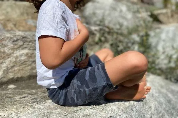 ein Kind sitze auf einem Stein im Schneidersitz und hält sich die Hände auf die Brust und den Bauch