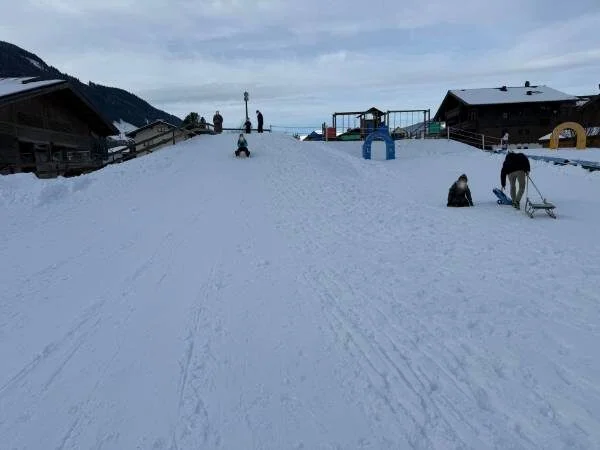 Kinder und Eltern genießen den Schneetag im Wintersportpark mit Schlittenfahren und Schneespielen, umgeben von Bergen und Winterhütten.
