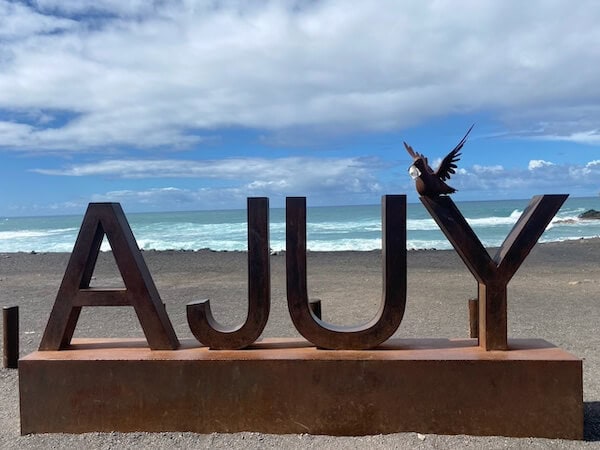 Der Blick auf den Strand mit gro&szlig;en Buchstaben "A J U Y" und einem Vogel, der auf dem Buchstaben sitzt, bei sonnigem Wetter und Wellen im Hintergrund, ideal f&uuml;r Familienurlaub auf Fuerteventura.