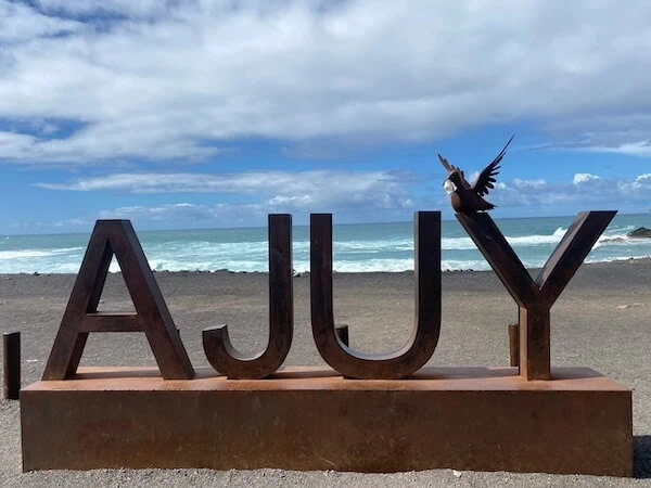 Der Blick auf den Strand mit großen Buchstaben "A J U Y" und einem Vogel, der auf dem Buchstaben sitzt, bei sonnigem Wetter und Wellen im Hintergrund, ideal für Familienurlaub auf Fuerteventura.