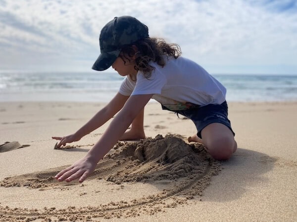 Ein junges M&auml;dchen baut im Sand am Strand auf Fuerteventura eine Sandburg, tr&auml;gt eine schwarze Cap und ein wei&szlig;es T-Shirt, ideal f&uuml;r Familienurlaub mit Kindern.