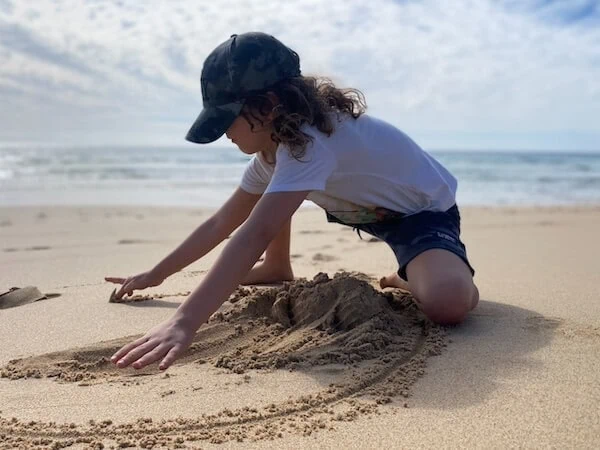 Ein junges Mädchen baut im Sand am Strand auf Fuerteventura eine Sandburg, trägt eine schwarze Cap und ein weißes T-Shirt, ideal für Familienurlaub mit Kindern.