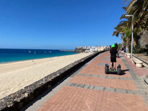 Fuerteventura mit Kindern: Familie beim Spaziergang entlang der Strandpromenade mit Segway, Palmen und Blick auf den Strand und das Meer, ideal f&uuml;r Familienurlaub mit Kindern.
