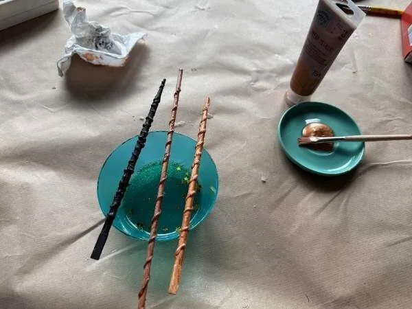 Fast fertige Zauberstäbe auf einem blauen Teller, kreative Bastelidee, dekorativ und kinderleicht, mit bereitgestellten Bastelmaterialien im Hintergrund.