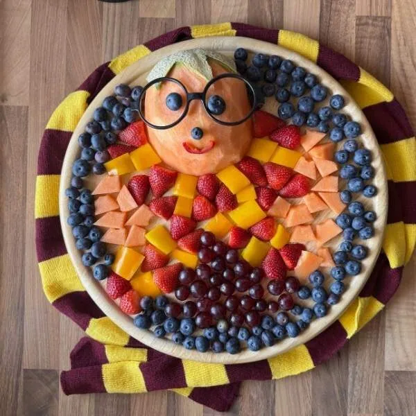 Obstteller mit frischem Obst, Paprika, als kreatives Harry Potter Obstgesicht und buntem Gemüsemotiv, liebevoll arrangiert auf einem große Teller mit buntem Stoff im Hintergrund.