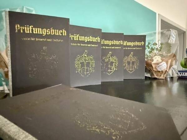 Prüfungsbücher aufgestellt nebeneinander und dahinter die Einladung zur Party mit der Alraune