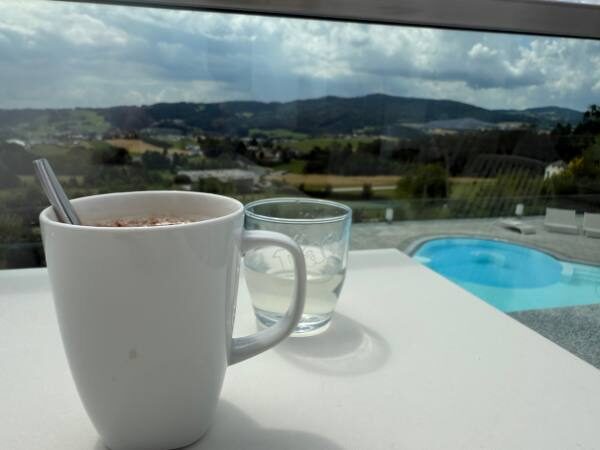 Im Vordergrund eine Tasse und im Hintergrund das Freibad und der Blick auf die Berge