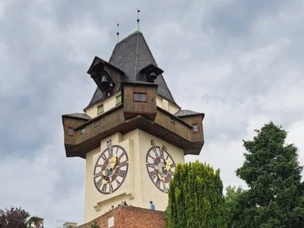 Historischer Uhrturm mit rustikalem Holzdesign und dicken Zifferblättern, umgeben von grünen Bäumen, auf einem bewölkten Himmel. Perfektes Motiv für Reisen, Geschichte und Architektur in Österreich.
