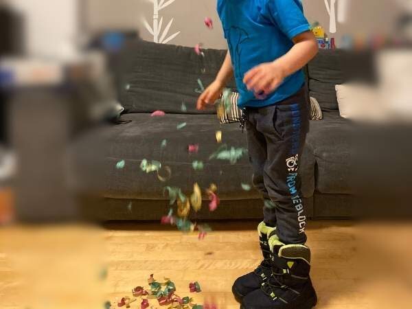 Bunte Legosteine fliegen durch die Luft, während ein Kind in Turnschuhen auf einem Teppich steht, im Wohnzimmer mit Spielzeug.