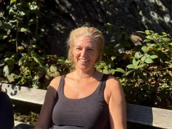 Lächelnde Frau im Sonnenlicht auf einer Parkbank, umgeben von grünen Pflanzen, entspannte Atmosphäre, natürlicher Hintergrund, sommerliche Freizeit, entspannte Momente, Outdoor-Aktivitäten, Wohlbefinden, Natur, aktive Lebensweise.