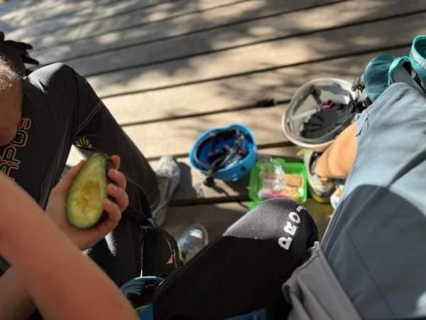Frühstückszeit am See, Eltern und Kinder genießen eine Snackpause während eines Familienausflugs, mit gesunden Früchten und Picknick-Zubehör an einem sonnigen Tag.