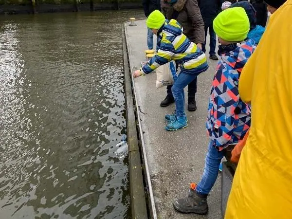 Kinder beim Experimentieren am Flussufer bei kaltem Wetter mit Regenjacken und Mützen.