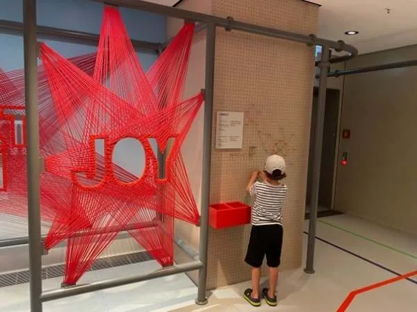 Farbenfrohes Inside-Design mit rotem "JOY" Schriftzug und einem Kind, das hinter einer Wand mit Pinnwand steht, ideal für Familien und Kinder im Indoor-Spielbereich.