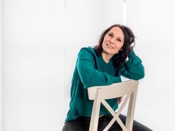 Entspannte Frau sitzt an einem weißen Stuhl vor weißer Wand, lächelt und freut sich.