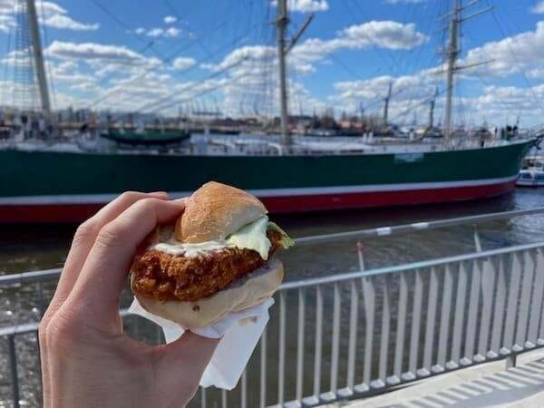 Eine Hand hält ein Fish Sandwich mit Fischfilet, Mayo und Brötchen vor einem Hafen mit Segelschiffen an einem sonnigen Tag.