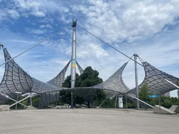 Moderne Skulptur aus Stahlkonstruktion vor blauem Himmel und Wolken in einem städtischen Park von München
