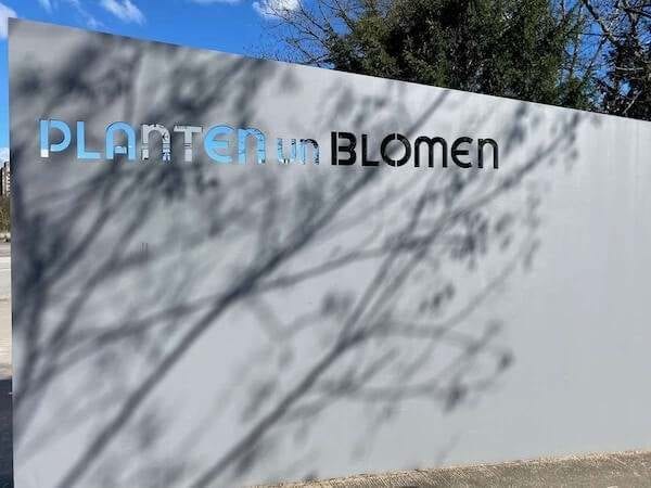 Schriftzug am Eingang vom Park in Hamburg "Planten u. Blomen" mit Schatten der Bäume im Hintergrund, modernes Design, Frühlingsblumen, Garten, Natur, Stadt, Blumentrends, Gartengestaltung, Ästhetik.