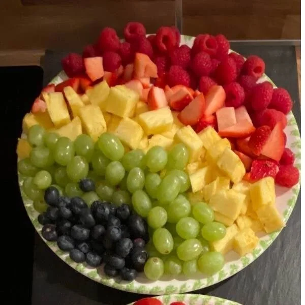 Bunte Regenbogen Obstplatte mit Weintrauben, Melone, Erdbeeren, Himbeeren und Ananas – gesunde Snacks für Kinder und Familie.