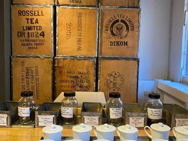 Hochwertige Teeproben in Gläsern auf einem Tisch, umgeben von Teekisten mit Marken wie Rossell Tea Limited, ideal für Tee-Enthusiasten und Genussliebhaber.