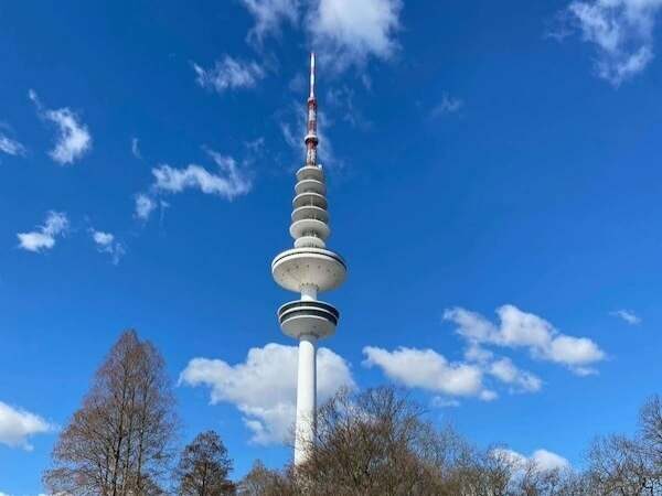Ein moderner Fernsehturm vor blauem Himmel mit Wolken, Umgeben von Bäumen, Symbol für Berliner Stadtbild, perfekte Kulisse für Familien und Touristen.
