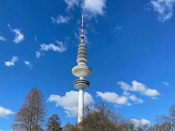 Ein moderner Fernsehturm vor blauem Himmel mit Wolken, Umgeben von Bäumen, Symbol für Berliner Stadtbild, perfekte Kulisse für Familien und Touristen.