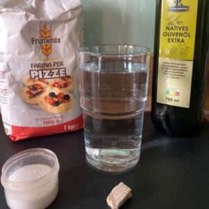 Zutaten für den Pizzateig auf einer Arbeitsplatte: Olivenölflasche, 1 Glas Wasser, Pizzamehl in der Packung, Salz in Plastikdoese und ein Stück Hefe