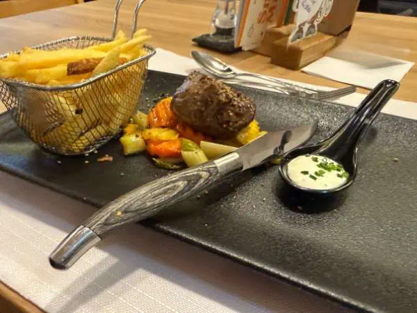 Zartes Rinderfilet mit Gemüse und knusprigen Pommes auf einem schwarzen Teller, serviert mit einer cremigen Sauce in einem kleinen Schälchen, ideal für Familienessen im Kinderhotel Felben.