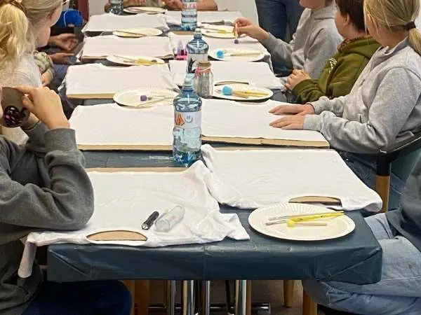 Kinder essen gemeinsam bei einer Veranstaltung, mit Wasserflaschen und Teller am Tisch.hirts und Pinseln