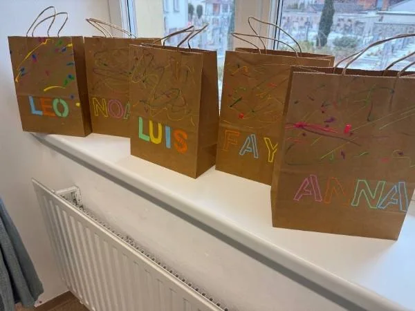 Mitgebseltüten für die Schwarzlichtparty mit dem Namenszug in Neon Schrift und einer bunten Glow in the Dark Farbspritzer stehen auf einem Fensterbrett 5 Stück