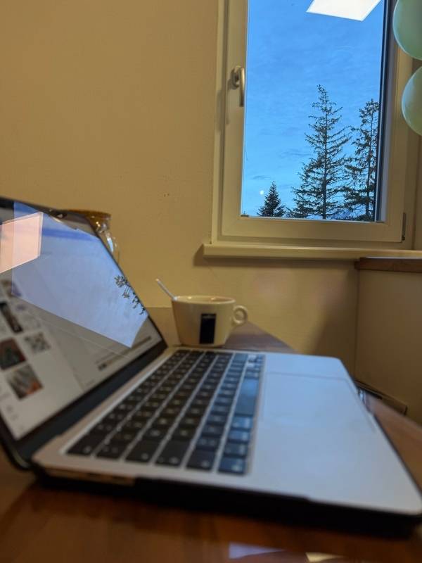 Laptop, Tasse und Fenster mit winterlichem Ausblick auf Tannenb&auml;ume, ideal f&uuml;r gem&uuml;tliche Wintertage und gegen Winterblues. Perfekt f&uuml;r eine entspannte Auszeit in der dunklen Jahreszeit.