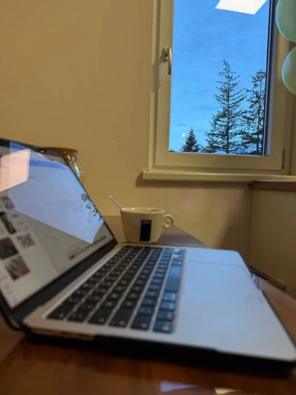 Laptop, Tasse und Fenster mit winterlichem Ausblick auf Tannenbäume, ideal für gemütliche Wintertage und gegen Winterblues. Perfekt für eine entspannte Auszeit in der dunklen Jahreszeit.
