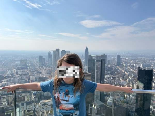 Frau mit ausgebreiteten Armen auf der Aussichtsplattform des Main Towers, Blick auf die Skyline von Frankfurt am Main an einem sonnigen Tag.