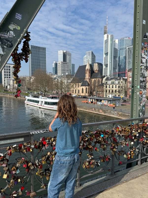Ein Mädchen mit braunen Haaren in Jeans und T-Shirt blickt auf die Skyline von Frankfurt am Main, während sie auf einer Brücke über den Fluss steht, umgeben von Liebesschlössern.