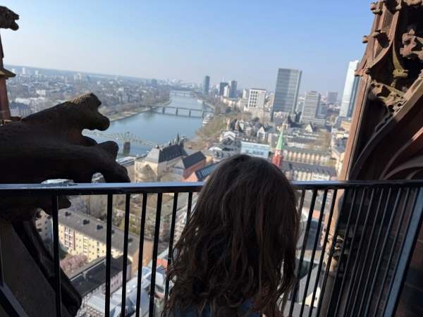 Hoch oben in einem Turm mit Blick auf die Stadt, eine Frau mit lockigem Haar genie&szlig;t die Aussicht auf Fluss und Skyline, umgeben von historischen Geb&auml;uden und moderner Architektur.
