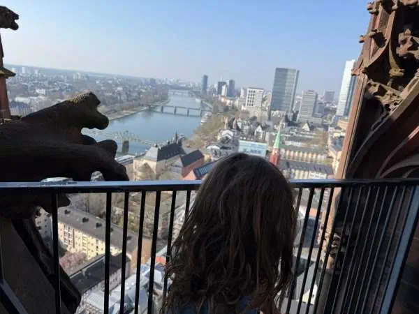 Hoch oben in einem Turm mit Blick auf die Stadt, eine Frau mit lockigem Haar genießt die Aussicht auf Fluss und Skyline, umgeben von historischen Gebäuden und moderner Architektur.