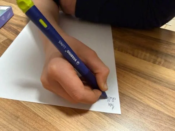 Ein Kind schreibt mit einem blauen Stabilo Lunis Ink Marker auf einem weißen Blatt Papier, das auf einem Holztisch liegt, im Rahmen des Lernens von Vokabeln.