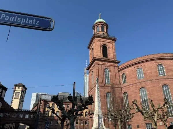 Ein rotes Backsteingebäude mit Kirchturm und blauer Himmel im Hintergrund, umgeben von geschnittenen Bäumen, in der Innenstadt, bei sonnigem Wetter, in Deutschland.