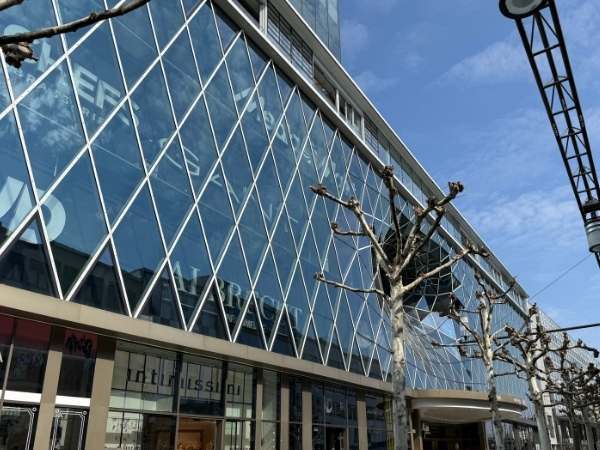 Einkaufszentrum mit moderner Glasfassade und Baum in der Stadt, sonniger Himmel, st&auml;dtisches Umfeld, urbanes Shopping-Erlebnis, klare Architektur, Stadtleben, moderne Infrastruktur, Stadtzentrum, urba.