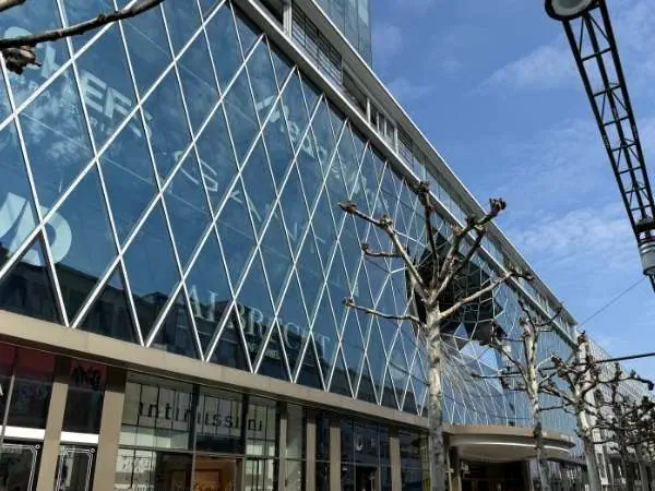 Einkaufszentrum mit moderner Glasfassade und Baum in der Stadt, sonniger Himmel, städtisches Umfeld, urbanes Shopping-Erlebnis, klare Architektur, Stadtleben, moderne Infrastruktur, Stadtzentrum, urba.