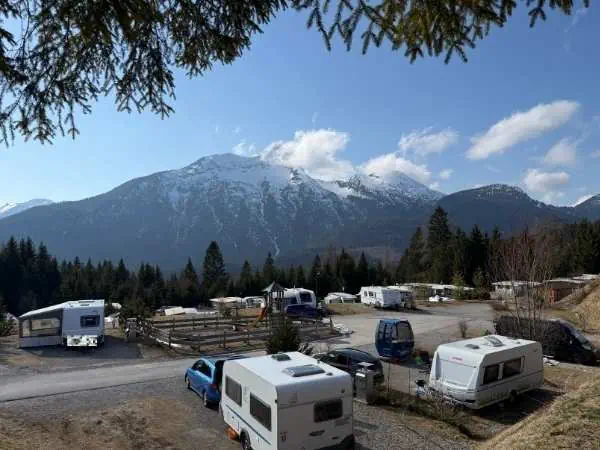 Bergpanorama mit schneebedecktem Gipfel, umgeben von Wäldern, mit Wohnmobilen und Autos auf einem Campingplatz im Vordergrund, bei schönem Wetter in den Bergen.