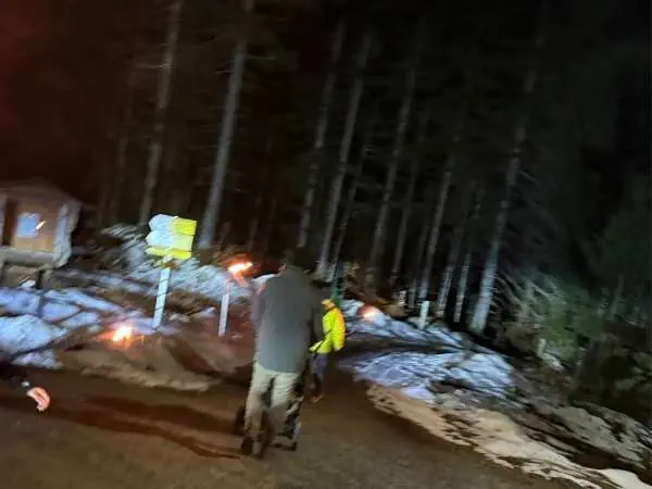 Ein Paar wandert nachts im verschneiten Wald, umgeben von Bäumen, bei dunklem Himmel, mit Taschenlampen und warmer Kleidung, in einer winterlichen, ruhigen Naturkulisse.