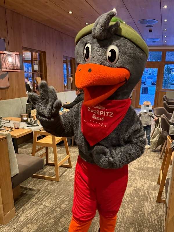 Ein Maskottchen in einem Restaurant, das einen Dinosaurier mit grüner Mütze und rotem Halstuch trägt, begrüßt Gäste und sorgt für Unterhaltung.
