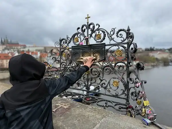 Ein Kind berührt ein kunstvolles, verziertes Gitter am Flussufer in Prag, Tschechien, bei bewölktem Himmel, während Familien die historische Stadt erkunden.