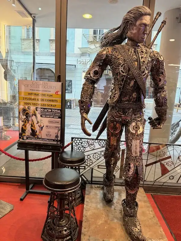 Detailreiche Skulptur eines Kriegers aus recyceltem Metall, präsentiert in einem Museum mit Sitzgelegenheiten im Vordergrund, ideal für Kunst- und Kulturinteressierte.