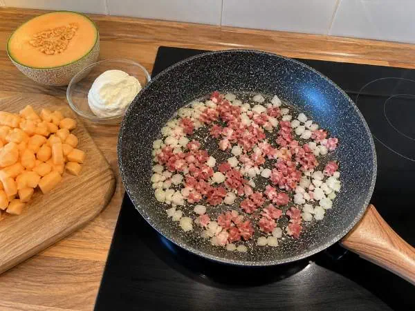 Zwiebeln und Speck werden in einer Pfanne angebraten, während auf einem Holzbrett gewürfelter Kürbis und eine Schüssel mit Sauerrahm bereitstehen.