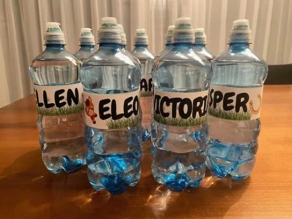 Mehrere Wasserflaschen mit individuell gestalteten Labels, die Namen wie Ellen, Eleonore, Victoria und Jasper tragen, auf einem Holztisch. Perfekt für personalisiertes Wasser bei Familien- oder Kinder.