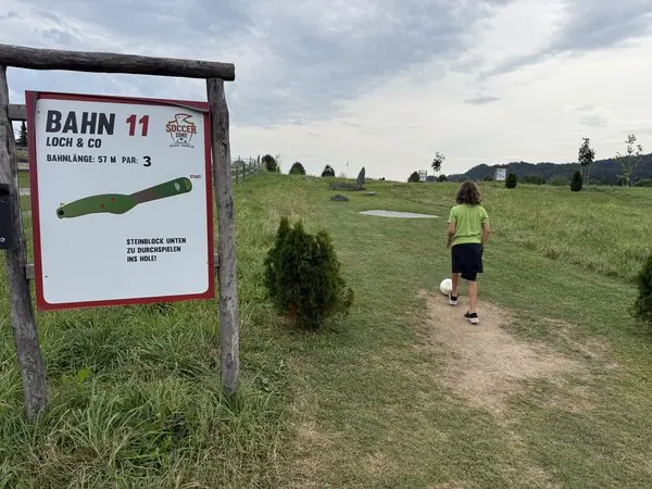 Junge beim Minigolf auf einer grünen Wiese, mit Bäumen und Himmel im Hintergrund, bei bewölktem Wetter, in einer ländlichen Umgebung, für MamaBlog optimiert.
