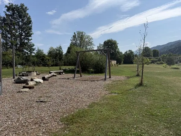 Ein Spielplatz im Freien mit Schaukel, Bäumen und Natur in einer ländlichen Umgebung, ideal für Kinder und Familien, bei schönem Wetter.