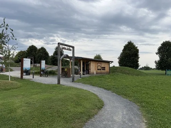 Ein Fahrradverleih- und Informationsstand in einem Park mit Fahrradwegen, Bäumen und Wolken am Himmel, ideal für Familien und Radfahrer in der Natur.